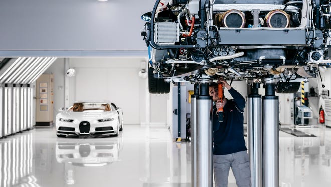 A Bugatti engineer works on the underside of a lifted Chiron, with a completed model sitting in the back of the room.